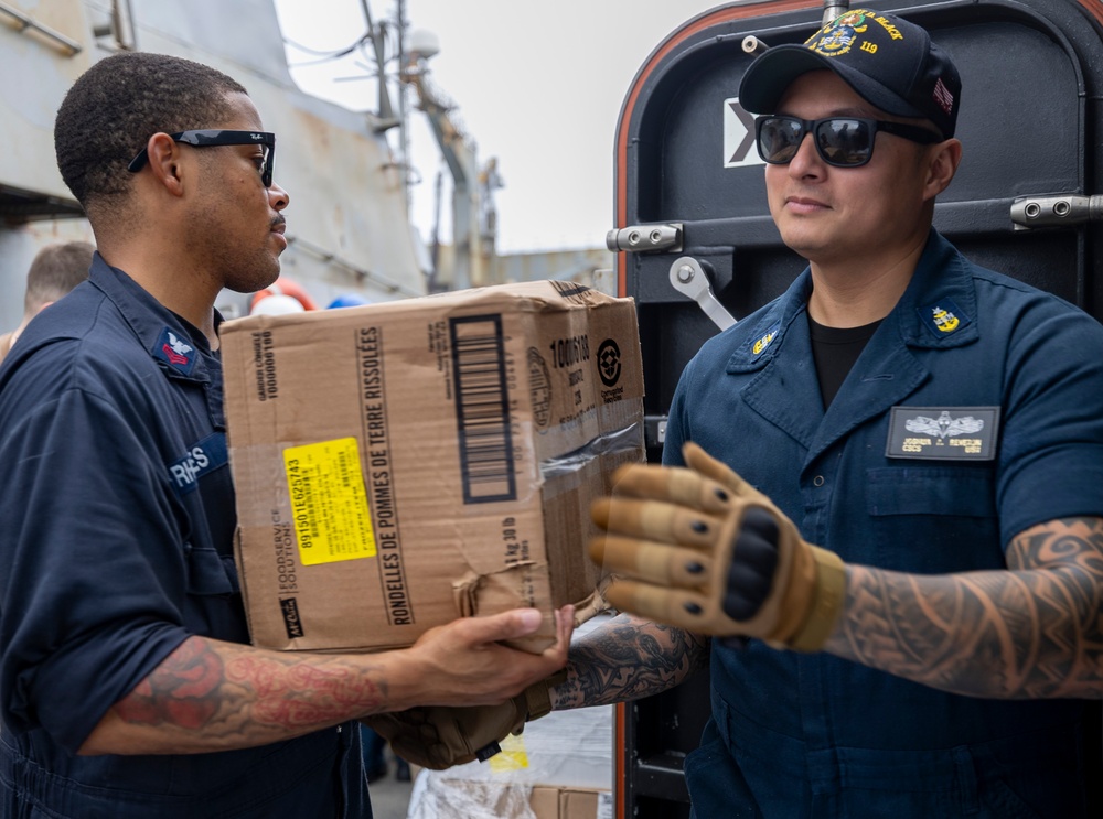 Two U.S. Navy sailors in dark blue uniforms are transferring a cardboard box labeled 'Rondelles de Pommes de Terre Rissolées' (hash brown potatoes) during what appears to be a replenishment or supply operation aboard a naval vessel. The sailor on the right wears a cap with '119' visible and tactical gloves, while both personnel display rank insignia and name tags. The setting appears to be an exterior deck area of a warship.