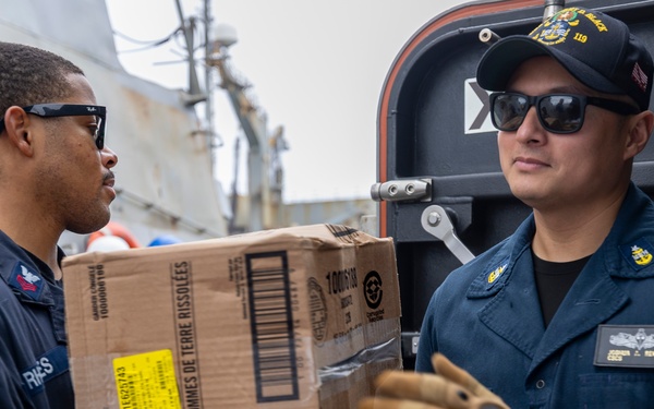 Two U.S. Navy sailors in dark blue uniforms are transferring a cardboard box labeled 'Rondelles de Pommes de Terre Rissolées' (hash brown potatoes) during what appears to be a replenishment or supply operation aboard a naval vessel. The sailor on the right wears a cap with '119' visible and tactical gloves, while both personnel display rank insignia and name tags. The setting appears to be an exterior deck area of a warship.