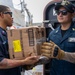 Two U.S. Navy sailors in dark blue uniforms are transferring a cardboard box labeled 'Rondelles de Pommes de Terre Rissolées' (hash brown potatoes) during what appears to be a replenishment or supply operation aboard a naval vessel. The sailor on the right wears a cap with '119' visible and tactical gloves, while both personnel display rank insignia and name tags. The setting appears to be an exterior deck area of a warship.