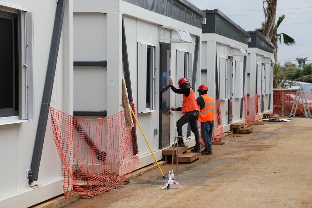 Two construction workers wearing orange high-visibility vests and red hard hats are working on a row of modular/prefabricated building units at an active construction site. The workers appear to be installing or adjusting a door on one of the units. The site is bordered by orange safety netting and shows signs of ongoing construction activity.