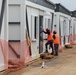 Two construction workers wearing orange high-visibility vests and red hard hats are working on a row of modular/prefabricated building units at an active construction site. The workers appear to be installing or adjusting a door on one of the units. The site is bordered by orange safety netting and shows signs of ongoing construction activity.
