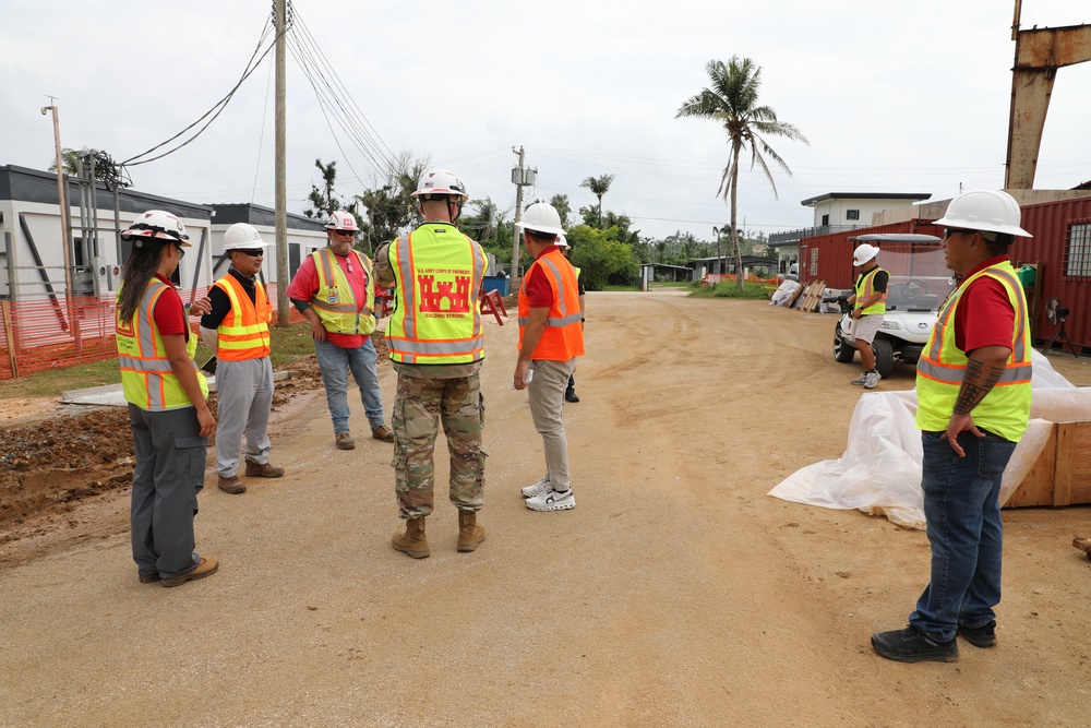 A group of construction and military personnel are gathered on a dirt road at an active construction site, appearing to hold a briefing or site walkthrough. One individual is wearing U.S. Army Corps of Engineers gear, identifiable by the castle emblem on their yellow safety vest. The setting appears to be a tropical location based on the palm trees and vegetation visible in the background.