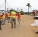 A group of construction and military personnel are gathered on a dirt road at an active construction site, appearing to hold a briefing or site walkthrough. One individual is wearing U.S. Army Corps of Engineers gear, identifiable by the castle emblem on their yellow safety vest. The setting appears to be a tropical location based on the palm trees and vegetation visible in the background.