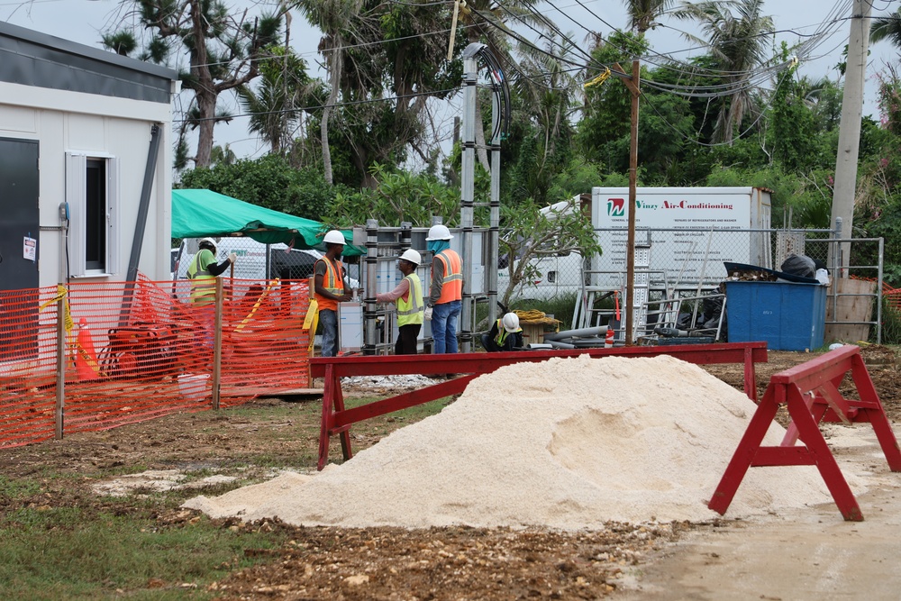 An active outdoor construction site featuring several workers in high-visibility safety gear and hard hats. The site includes a temporary office trailer, orange safety fencing, a pile of sand or fine aggregate, and various construction materials. A sign for 'Vinzy Air-Conditioning' is visible in the background near a chain-link fence.