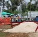 An active outdoor construction site featuring several workers in high-visibility safety gear and hard hats. The site includes a temporary office trailer, orange safety fencing, a pile of sand or fine aggregate, and various construction materials. A sign for 'Vinzy Air-Conditioning' is visible in the background near a chain-link fence.