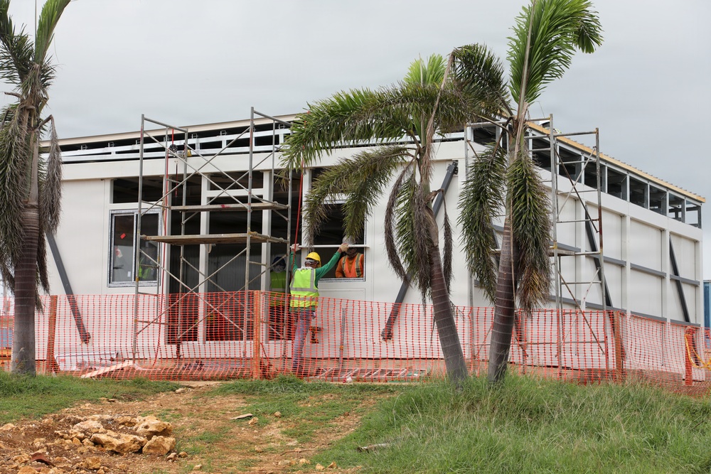 A building under active construction is shown with scaffolding erected along its exterior walls. Two workers in high-visibility safety gear are visible installing or adjusting a window panel on the facade. The site is surrounded by orange safety fencing, with palm trees in the foreground and an overcast sky above.