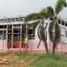 A building under active construction is shown with scaffolding erected along its exterior walls. Two workers in high-visibility safety gear are visible installing or adjusting a window panel on the facade. The site is surrounded by orange safety fencing, with palm trees in the foreground and an overcast sky above.