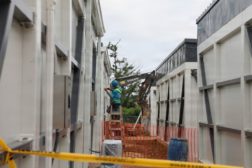 A construction worker on a ladder is performing work on the exterior wall of a modular or prefabricated structure in a narrow corridor between two large white panel units. A Volvo excavator is visible in the background operating within the same confined space. The site is cordoned off with orange safety fencing and yellow caution tape in the foreground.