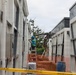 A construction worker on a ladder is performing work on the exterior wall of a modular or prefabricated structure in a narrow corridor between two large white panel units. A Volvo excavator is visible in the background operating within the same confined space. The site is cordoned off with orange safety fencing and yellow caution tape in the foreground.