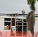 A construction site showing workers actively engaged in building work on a two-story modular or prefabricated structure. At least two workers are visible — one on the rooftop wearing a high-visibility vest and hard hat, and another at ground/lower level in an orange vest and white hard hat. The site is secured with orange safety fencing and scaffolding, with a palm tree visible to the right.