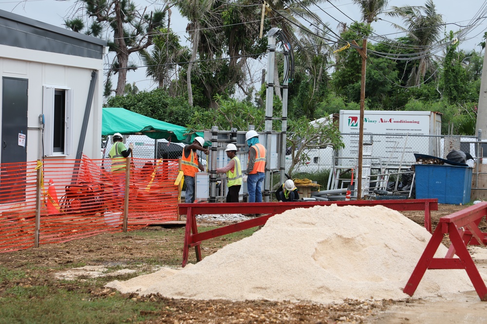 An active outdoor construction site featuring multiple workers in high-visibility safety vests and hard hats. The site includes temporary structures, safety barriers, and construction materials. A white truck with 'Winzy Air-Conditioning' branding is visible in the background.