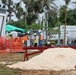 An active outdoor construction site featuring multiple workers in high-visibility safety vests and hard hats. The site includes temporary structures, safety barriers, and construction materials. A white truck with 'Winzy Air-Conditioning' branding is visible in the background.