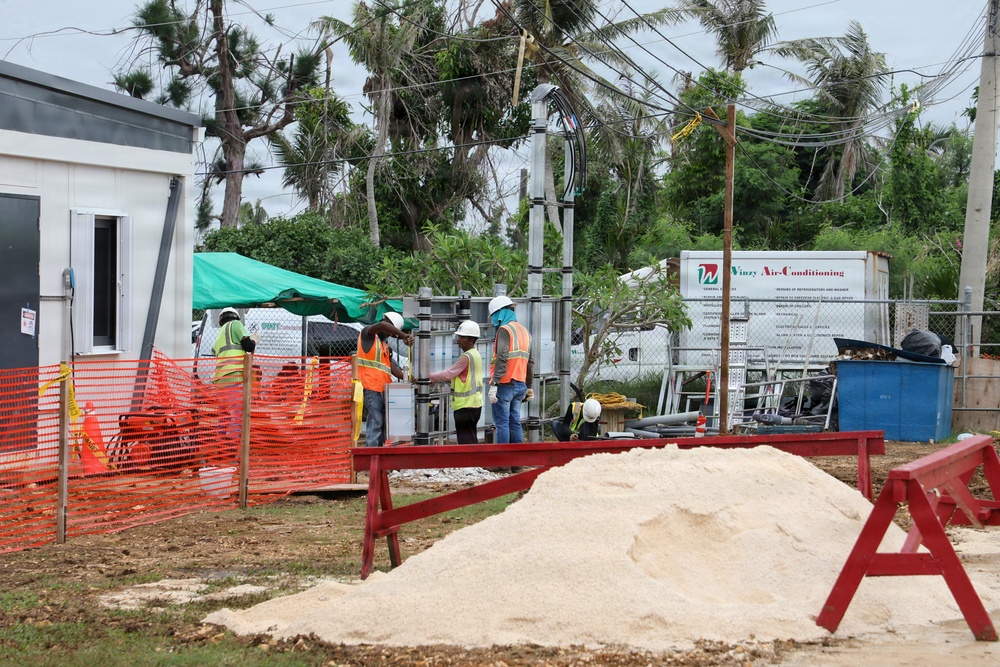 A construction site showing several workers in high-visibility safety vests and white hard hats working around equipment and materials. The site features orange safety fencing, a pile of sand or aggregate material in the foreground, and a truck branded 'Vinzy Air-Conditioning' visible in the background. The surrounding trees appear storm-damaged, suggesting this may be post-hurricane recovery or reconstruction work.