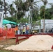 A construction site showing several workers in high-visibility safety vests and white hard hats working around equipment and materials. The site features orange safety fencing, a pile of sand or aggregate material in the foreground, and a truck branded 'Vinzy Air-Conditioning' visible in the background. The surrounding trees appear storm-damaged, suggesting this may be post-hurricane recovery or reconstruction work.