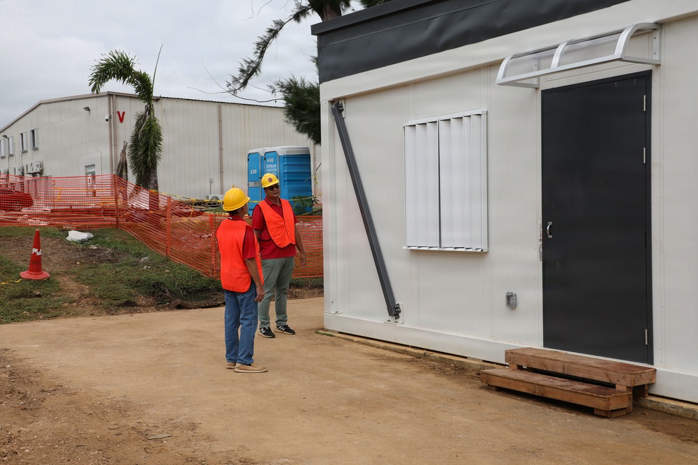 Two construction workers wearing orange safety vests and yellow hard hats are conversing near a modular/prefabricated white building unit at an active construction site. The site features orange safety fencing, a portable toilet, traffic cones, and a larger warehouse-style building visible in the background. The setting appears to be a tropical or subtropical location based on the palm trees and vegetation.