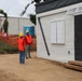 Two construction workers wearing orange safety vests and yellow hard hats are conversing near a modular/prefabricated white building unit at an active construction site. The site features orange safety fencing, a portable toilet, traffic cones, and a larger warehouse-style building visible in the background. The setting appears to be a tropical or subtropical location based on the palm trees and vegetation.