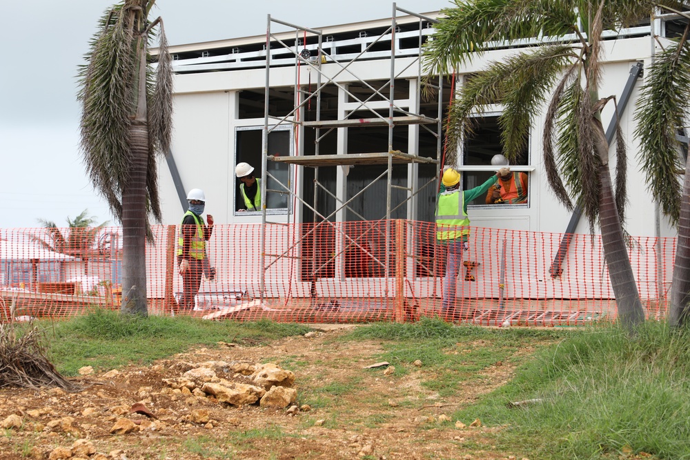 A construction site showing workers actively engaged in building or renovating a structure. Three workers wearing high-visibility vests and hard hats are visible, with scaffolding erected against the building facade. The site is cordoned off with orange safety netting, and palm trees are present in the foreground.