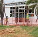 A construction site showing workers actively engaged in building or renovating a structure. Three workers wearing high-visibility vests and hard hats are visible, with scaffolding erected against the building facade. The site is cordoned off with orange safety netting, and palm trees are present in the foreground.