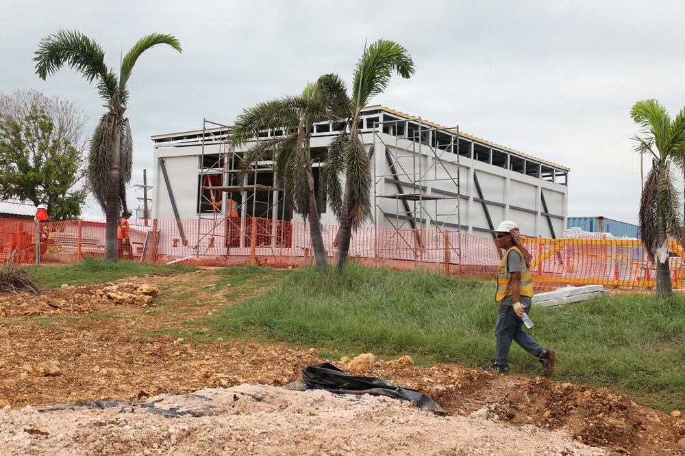 A construction site featuring a partially built steel-frame commercial or industrial building surrounded by palm trees, suggesting a tropical location. A worker in safety gear walks across disturbed ground in the foreground, while scaffolding and orange safety fencing are visible around the structure. The site shows active construction with exposed soil, rocky debris, and multiple workers present.