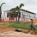 A construction site featuring a partially built steel-frame commercial or industrial building surrounded by palm trees, suggesting a tropical location. A worker in safety gear walks across disturbed ground in the foreground, while scaffolding and orange safety fencing are visible around the structure. The site shows active construction with exposed soil, rocky debris, and multiple workers present.