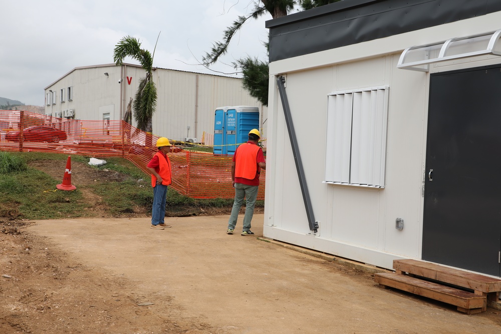 A construction site showing two workers wearing orange safety vests and yellow hard hats near a portable modular office building. The site features safety fencing, a traffic cone, portable toilets, and a large warehouse structure in the background. The setting appears to be in a tropical or subtropical location based on the palm trees visible.