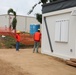 A construction site showing two workers wearing orange safety vests and yellow hard hats near a portable modular office building. The site features safety fencing, a traffic cone, portable toilets, and a large warehouse structure in the background. The setting appears to be in a tropical or subtropical location based on the palm trees visible.