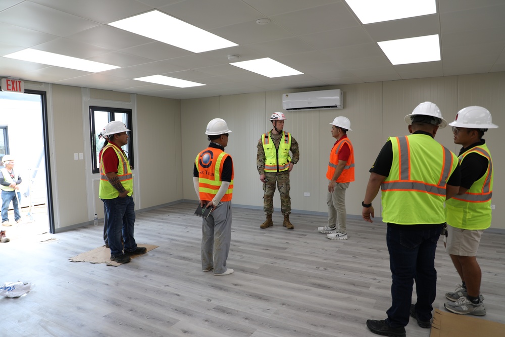 A group of approximately seven individuals are gathered inside a newly constructed or recently completed modular building interior. One person in military camouflage uniform and a red-marked hard hat appears to be briefing or addressing the group, who are wearing high-visibility safety vests and white hard hats. The space features finished flooring, recessed LED lighting, a wall-mounted mini-split HVAC unit, and an exit sign near an open door.