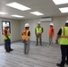 A group of approximately seven individuals are gathered inside a newly constructed or recently completed modular building interior. One person in military camouflage uniform and a red-marked hard hat appears to be briefing or addressing the group, who are wearing high-visibility safety vests and white hard hats. The space features finished flooring, recessed LED lighting, a wall-mounted mini-split HVAC unit, and an exit sign near an open door.