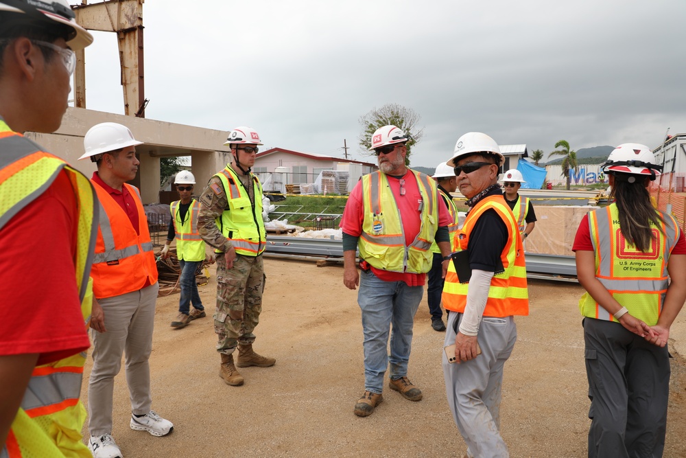 A group of construction and military personnel are gathered at an active construction site for what appears to be a briefing or site walkthrough. The group includes U.S. Army Corps of Engineers personnel, a soldier in camouflage uniform, and several workers wearing high-visibility safety vests and white hard hats. The setting shows a partially demolished or under-construction building in a tropical environment.