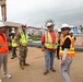 A group of construction and military personnel are gathered at an active construction site for what appears to be a briefing or site walkthrough. The group includes U.S. Army Corps of Engineers personnel, a soldier in camouflage uniform, and several workers wearing high-visibility safety vests and white hard hats. The setting shows a partially demolished or under-construction building in a tropical environment.