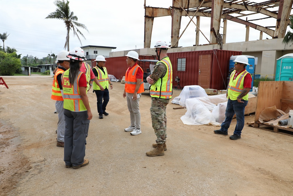 A group of approximately eight individuals wearing hard hats and high-visibility safety vests are gathered at an active construction or demolition site. One person appears to be in military camouflage uniform, while others wear civilian clothing with safety gear bearing what appears to be U.S. Army Corps of Engineers markings. The site features a partially demolished or damaged steel-frame structure, shipping containers, and construction materials in a tropical setting.