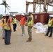 A group of approximately eight individuals wearing hard hats and high-visibility safety vests are gathered at an active construction or demolition site. One person appears to be in military camouflage uniform, while others wear civilian clothing with safety gear bearing what appears to be U.S. Army Corps of Engineers markings. The site features a partially demolished or damaged steel-frame structure, shipping containers, and construction materials in a tropical setting.