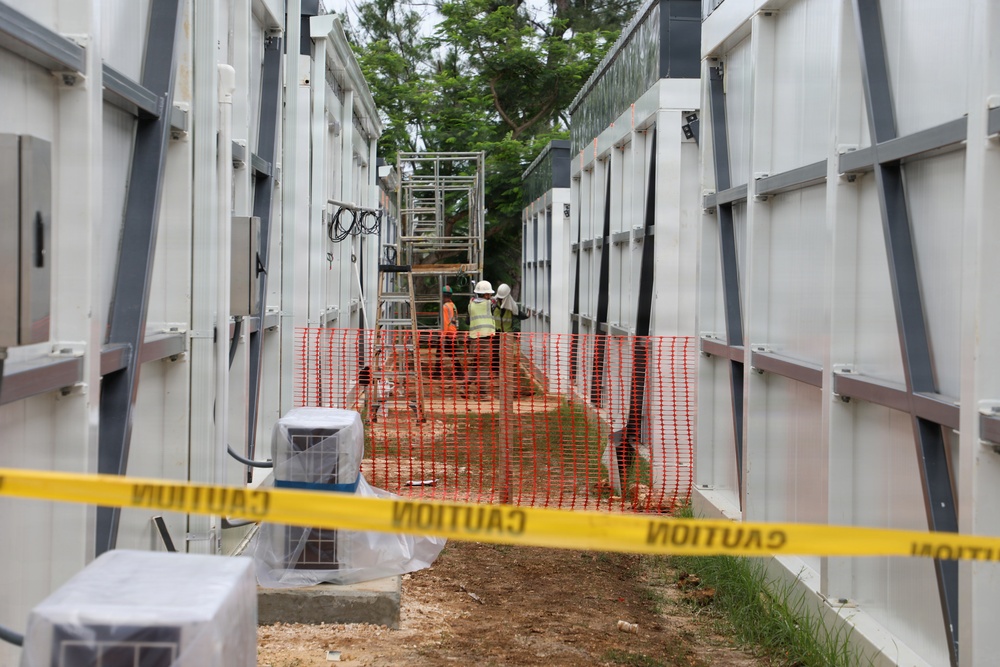 The image shows an active construction or installation site featuring rows of large white modular units or containers arranged in parallel corridors. Workers in high-visibility safety gear are visible mid-ground, working near a metal scaffold or ladder structure. The area is cordoned off with both yellow caution tape in the foreground and orange safety fencing further back.