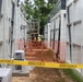 The image shows an active construction or installation site featuring rows of large white modular units or containers arranged in parallel corridors. Workers in high-visibility safety gear are visible mid-ground, working near a metal scaffold or ladder structure. The area is cordoned off with both yellow caution tape in the foreground and orange safety fencing further back.