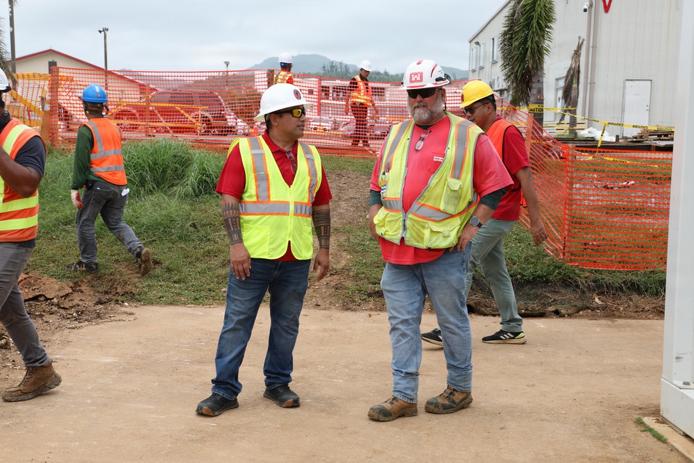 A construction site scene showing multiple workers in personal protective equipment including high-visibility vests and hard hats. Two workers in the foreground appear to be in conversation, while others are active in the background near orange safety fencing. The setting appears to be an outdoor construction or disaster recovery site with temporary structures visible.
