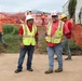 A construction site scene showing multiple workers in personal protective equipment including high-visibility vests and hard hats. Two workers in the foreground appear to be in conversation, while others are active in the background near orange safety fencing. The setting appears to be an outdoor construction or disaster recovery site with temporary structures visible.