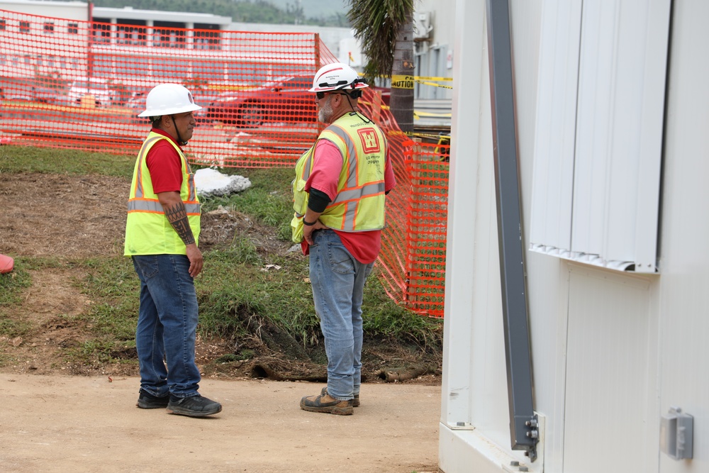 Two construction workers wearing high-visibility yellow safety vests and white hard hats are standing on a dirt path at an active construction site. They appear to be engaged in a conversation or discussion, possibly reviewing site conditions. The background shows orange safety fencing, caution tape, and a red building, indicating an active work zone.