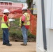 Two construction workers wearing high-visibility yellow safety vests and white hard hats are standing on a dirt path at an active construction site. They appear to be engaged in a conversation or discussion, possibly reviewing site conditions. The background shows orange safety fencing, caution tape, and a red building, indicating an active work zone.