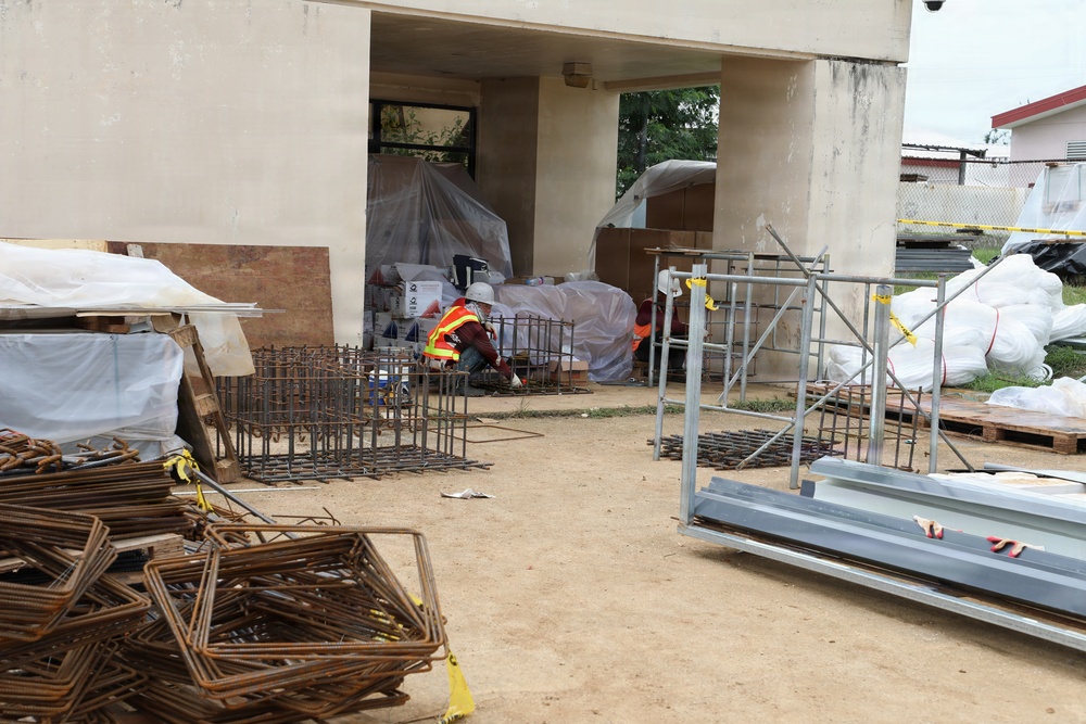 An active construction or renovation site adjacent to a concrete building, with various building materials and equipment staged in an outdoor area. A single worker wearing high-visibility orange safety vest and white hard hat is visible, crouching near rebar cage assemblies. The site contains a mix of steel reinforcement materials, scaffolding components, and covered supplies.
