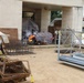 An active construction or renovation site adjacent to a concrete building, with various building materials and equipment staged in an outdoor area. A single worker wearing high-visibility orange safety vest and white hard hat is visible, crouching near rebar cage assemblies. The site contains a mix of steel reinforcement materials, scaffolding components, and covered supplies.