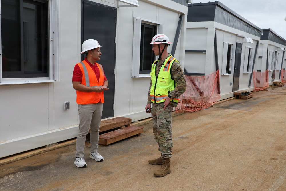 Two individuals are conversing at an active construction or temporary facility site. One person wears civilian attire with an orange safety vest and white hard hat, while the other is dressed in military camouflage uniform with a yellow safety vest and white hard hat. They are standing in front of a row of modular/prefabricated white buildings.