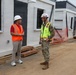 Two individuals are conversing at an active construction or temporary facility site. One person wears civilian attire with an orange safety vest and white hard hat, while the other is dressed in military camouflage uniform with a yellow safety vest and white hard hat. They are standing in front of a row of modular/prefabricated white buildings.