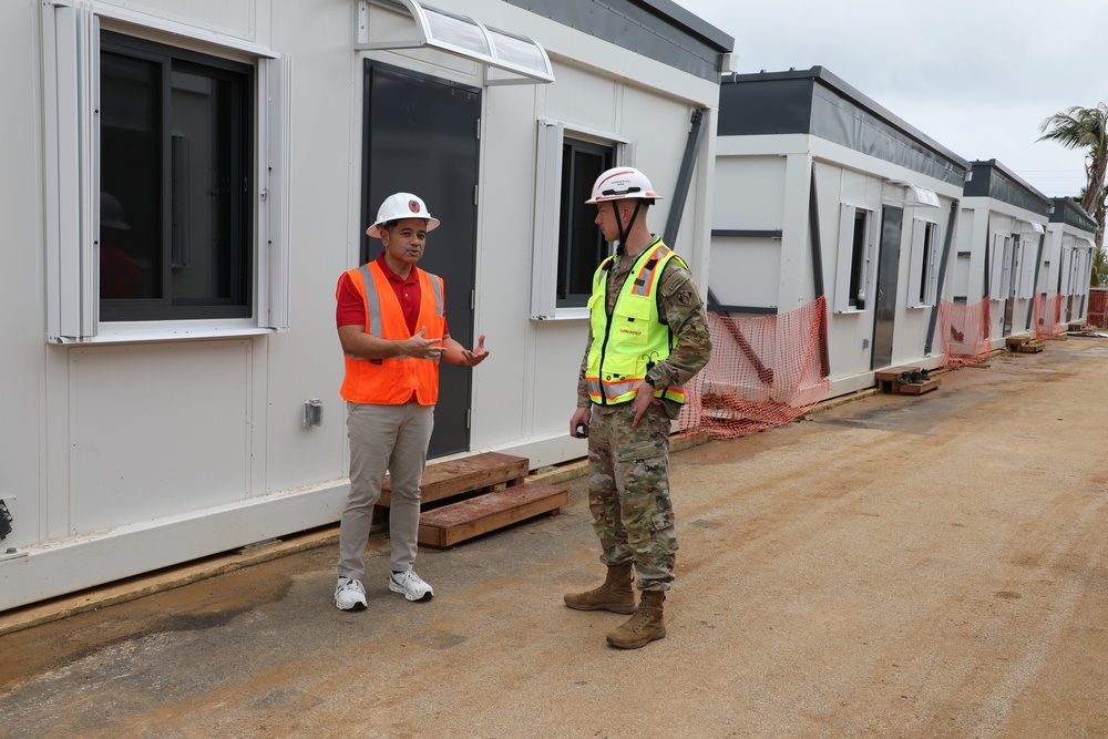 Two individuals are conversing at an active construction or installation site featuring a row of modular/prefabricated buildings. One person is wearing civilian construction attire with an orange safety vest, while the other is in military camouflage uniform with a yellow safety vest. The setting appears to be an outdoor area with dirt ground, orange safety fencing, and temporary modular structures extending into the background.