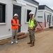 Two individuals are conversing at an active construction or installation site featuring a row of modular/prefabricated buildings. One person is wearing civilian construction attire with an orange safety vest, while the other is in military camouflage uniform with a yellow safety vest. The setting appears to be an outdoor area with dirt ground, orange safety fencing, and temporary modular structures extending into the background.