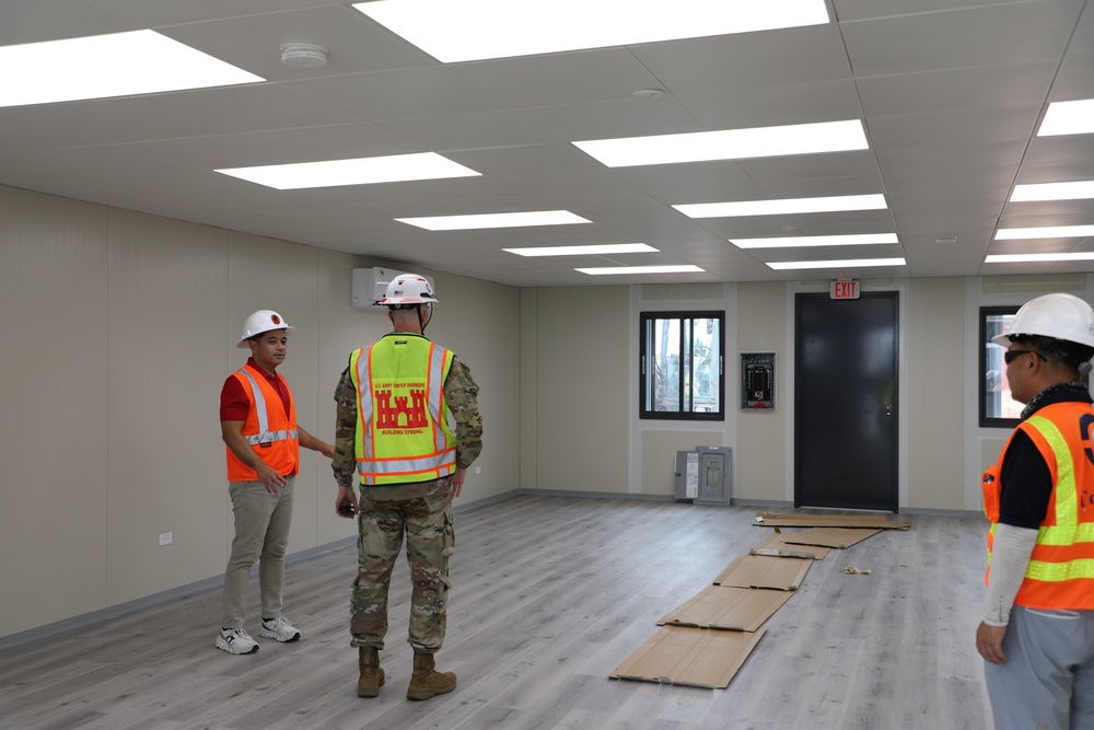 Three individuals are conducting an inspection inside a newly constructed or recently completed modular building interior. Two of the individuals are wearing high-visibility safety vests and white hard hats, while one is dressed in military camouflage uniform with a yellow safety vest bearing U.S. Army Corps of Engineers insignia. The space appears to be in a near-complete state with finished flooring, ceiling panels, and lighting installed.