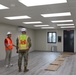 Three individuals are conducting an inspection inside a newly constructed or recently completed modular building interior. Two of the individuals are wearing high-visibility safety vests and white hard hats, while one is dressed in military camouflage uniform with a yellow safety vest bearing U.S. Army Corps of Engineers insignia. The space appears to be in a near-complete state with finished flooring, ceiling panels, and lighting installed.