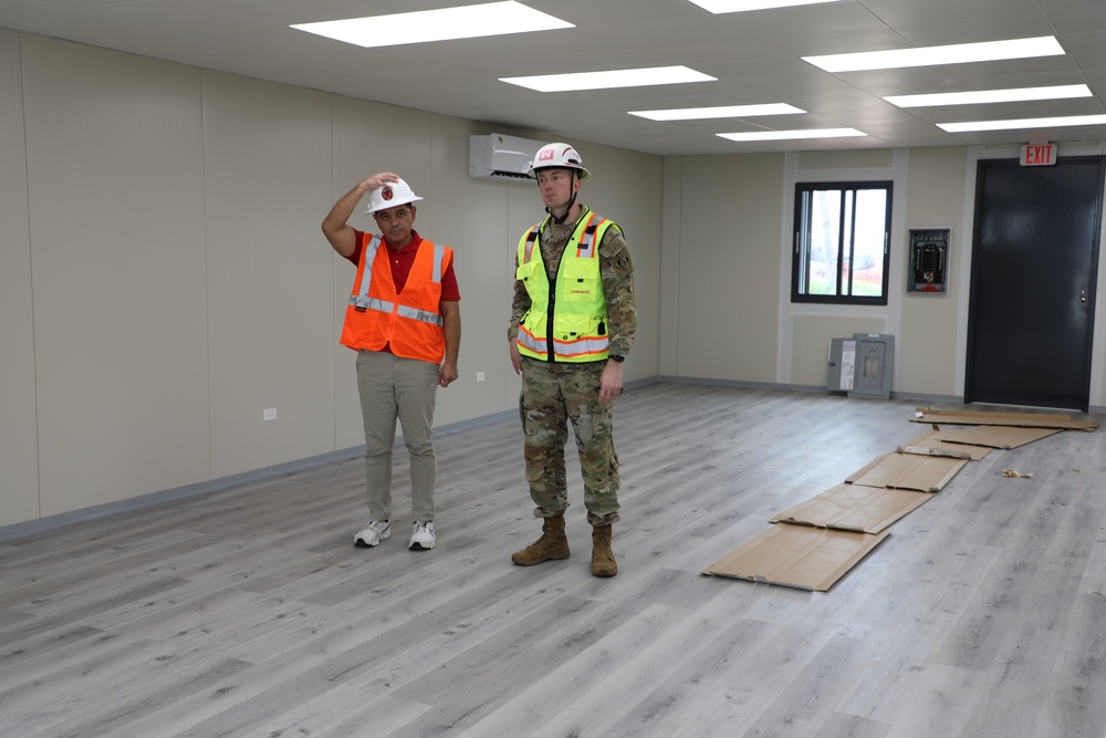 Two individuals stand inside a newly constructed or renovated interior space. One person wears an orange safety vest and white hard hat, while the other is dressed in military camouflage uniform with a yellow safety vest and white hard hat. The room appears to be in the final stages of completion, with a clean vinyl plank floor and cardboard protective covering still laid out.
