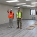 Two individuals stand inside a newly constructed or renovated interior space. One person wears an orange safety vest and white hard hat, while the other is dressed in military camouflage uniform with a yellow safety vest and white hard hat. The room appears to be in the final stages of completion, with a clean vinyl plank floor and cardboard protective covering still laid out.