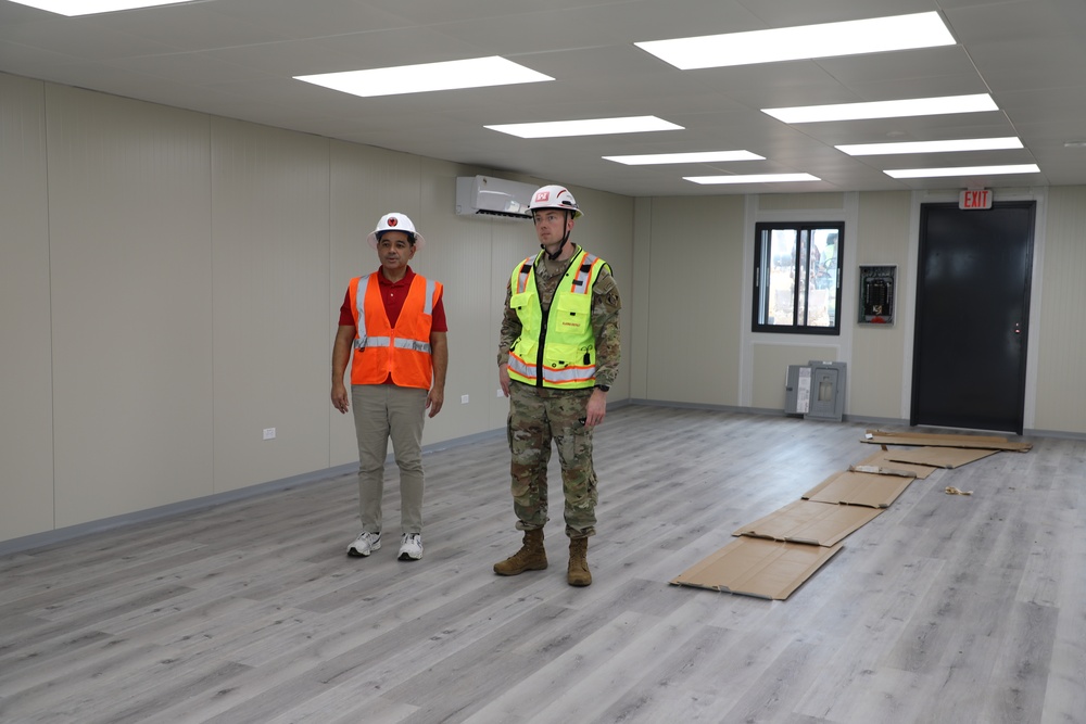 Two individuals stand inside a newly constructed or recently completed interior space with modern flooring and lighting. One person wears civilian construction attire with an orange safety vest, while the other is dressed in military camouflage uniform with a yellow safety vest. The room appears to be in the final stages of completion, with cardboard floor protection still laid out.
