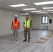 Two individuals stand inside a newly constructed or recently completed interior space with modern flooring and lighting. One person wears civilian construction attire with an orange safety vest, while the other is dressed in military camouflage uniform with a yellow safety vest. The room appears to be in the final stages of completion, with cardboard floor protection still laid out.